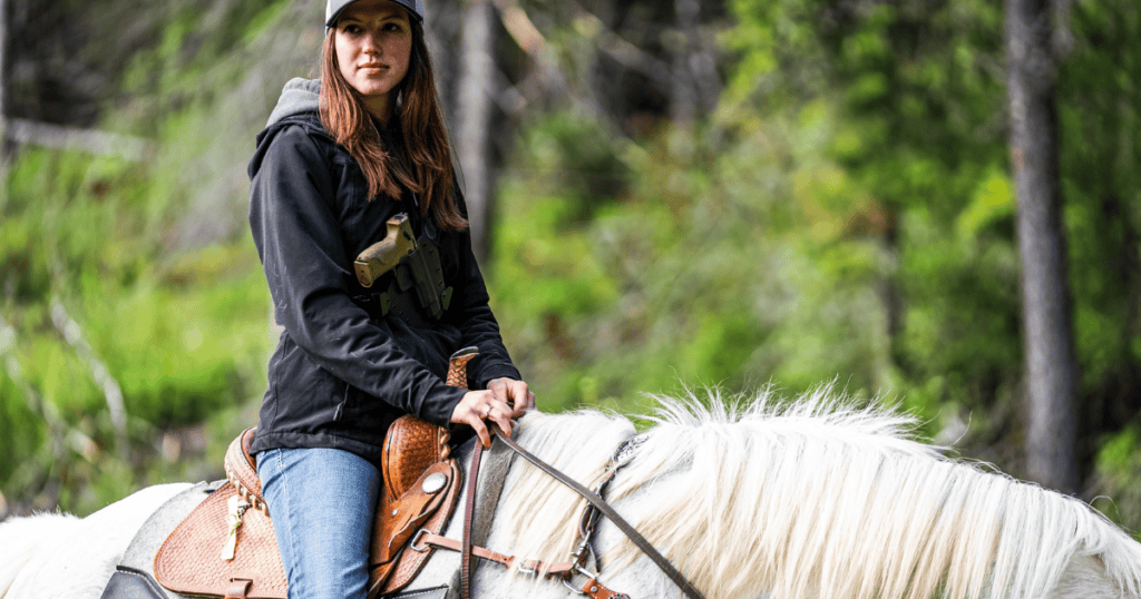 A young woman riding a horse carrying a gun with a chest holster by GunfightersINC.