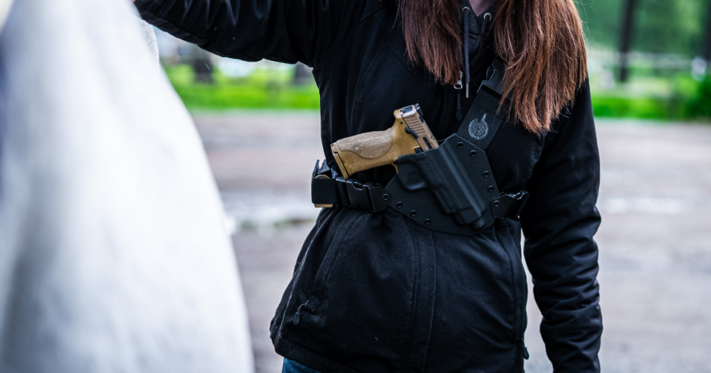 A woman standing next to a white horse wearing the Kenai Chest Holster by GunfightersINC.