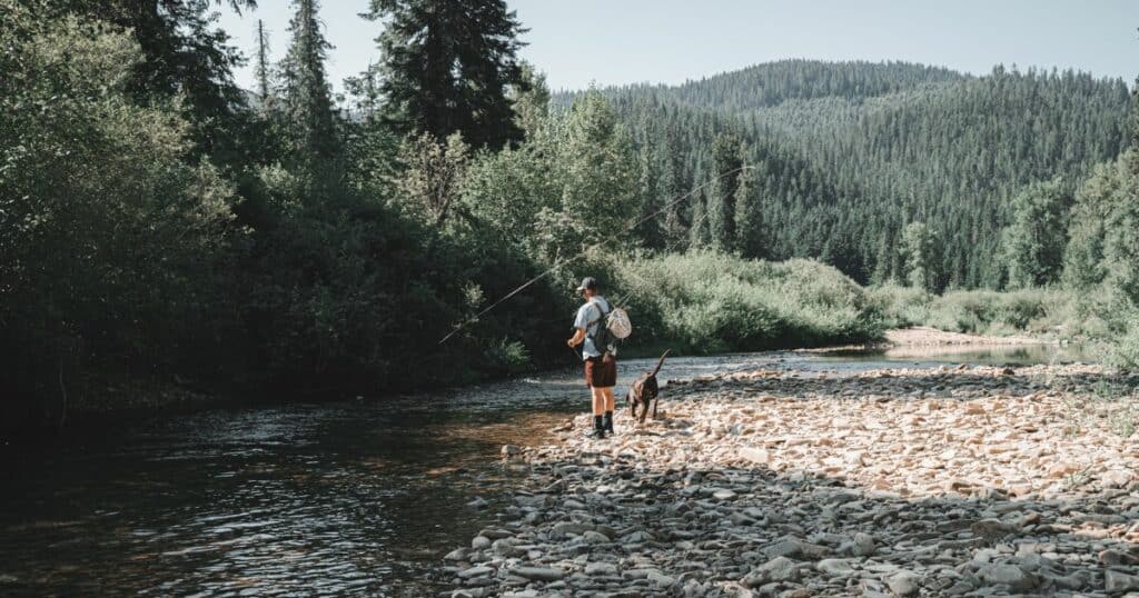 A man is fishing on a river with his dog while carrying a gun.