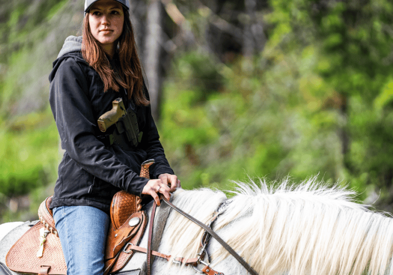 A young woman riding a horse carrying a gun with a chest holster by GunfightersINC.