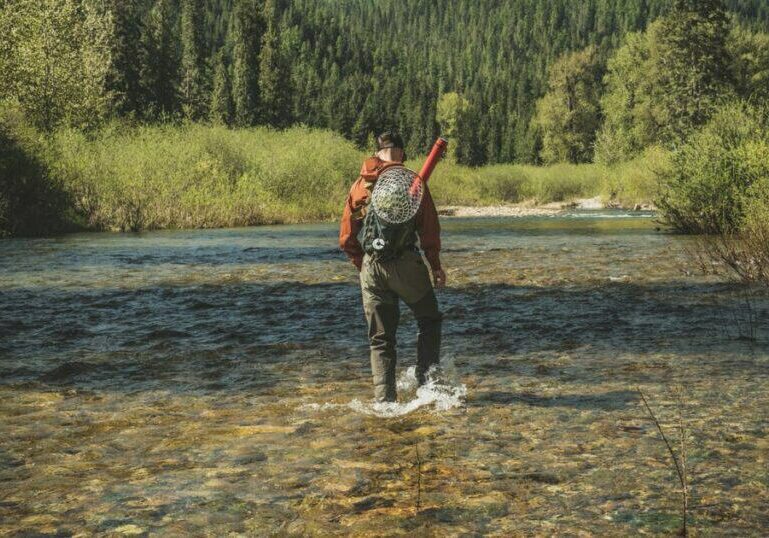 A man wearing waders walking through a shallow river to go fly fishing.