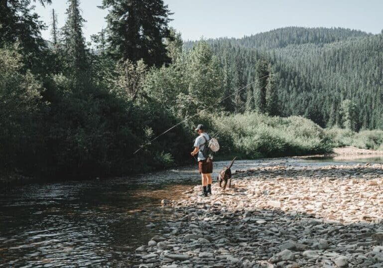 A man is fishing on a river with his dog while carrying a gun.