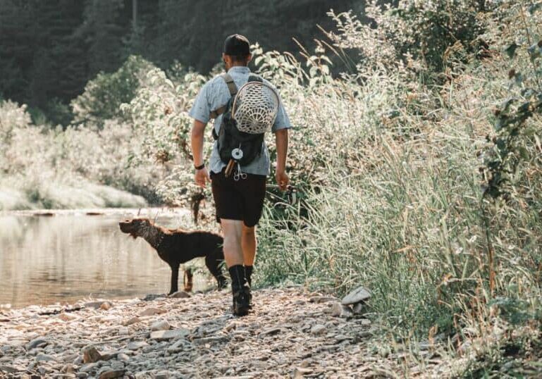 A man walking along a river with his dog looking for a fishing spot.